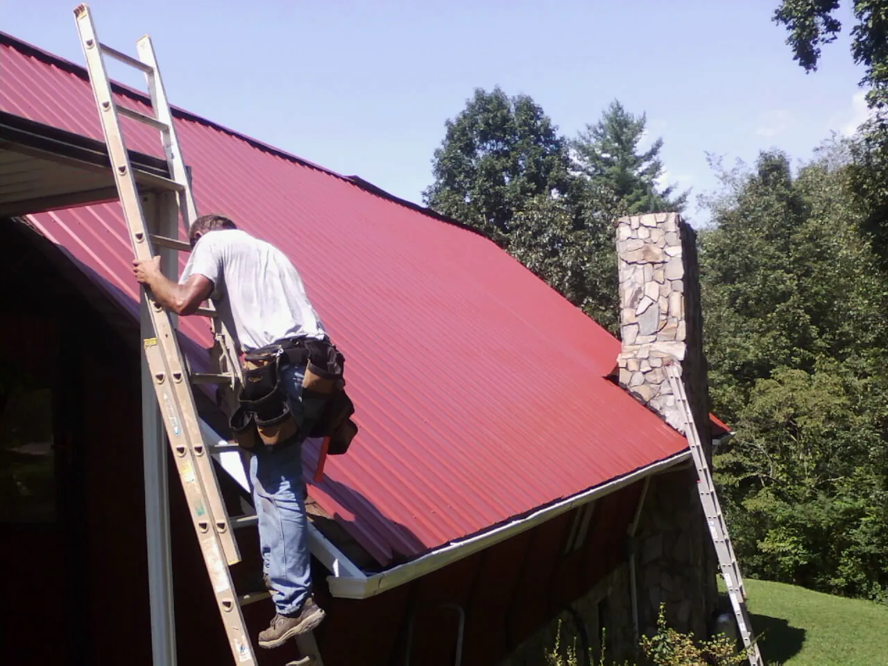 Roofer on a ladder beside a red metal roof and stone chimney — Soffit & Fascia Repair by Roofing Brothers in Syosset