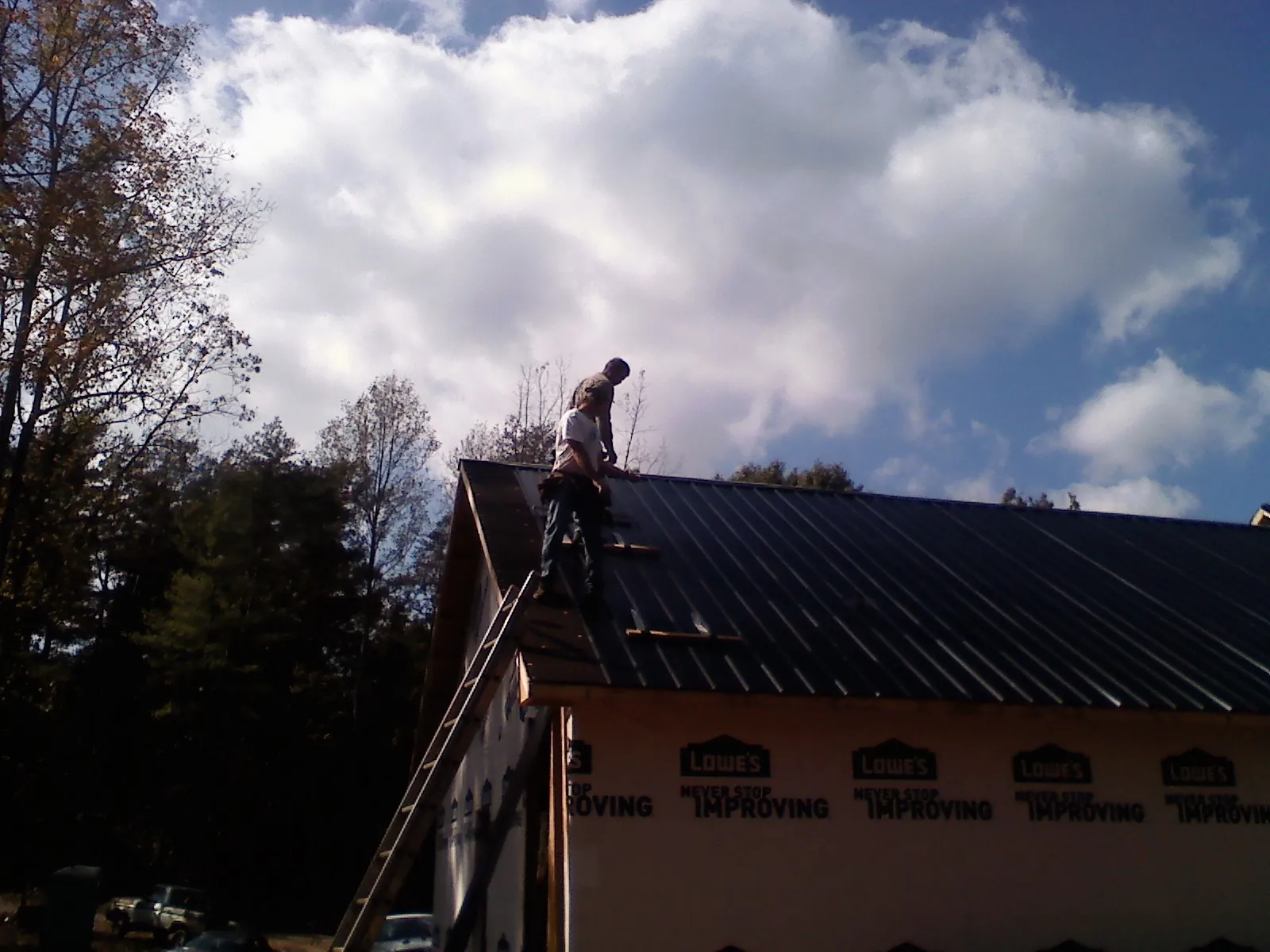 Roofer fastening dark metal panels high against a cloudy sky, from a Roof Inspection job in Syosset